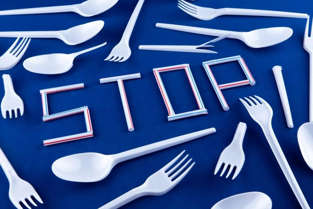 The word "STOP" spelled out using plastic straws, surrounded by various plastic utensils on a blue background.