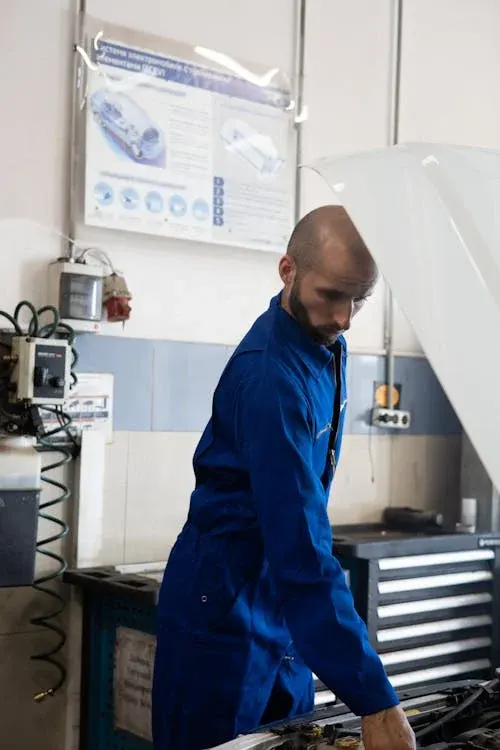 A mechanic in a blue jumpsuit working under the hood of a car in a garage.