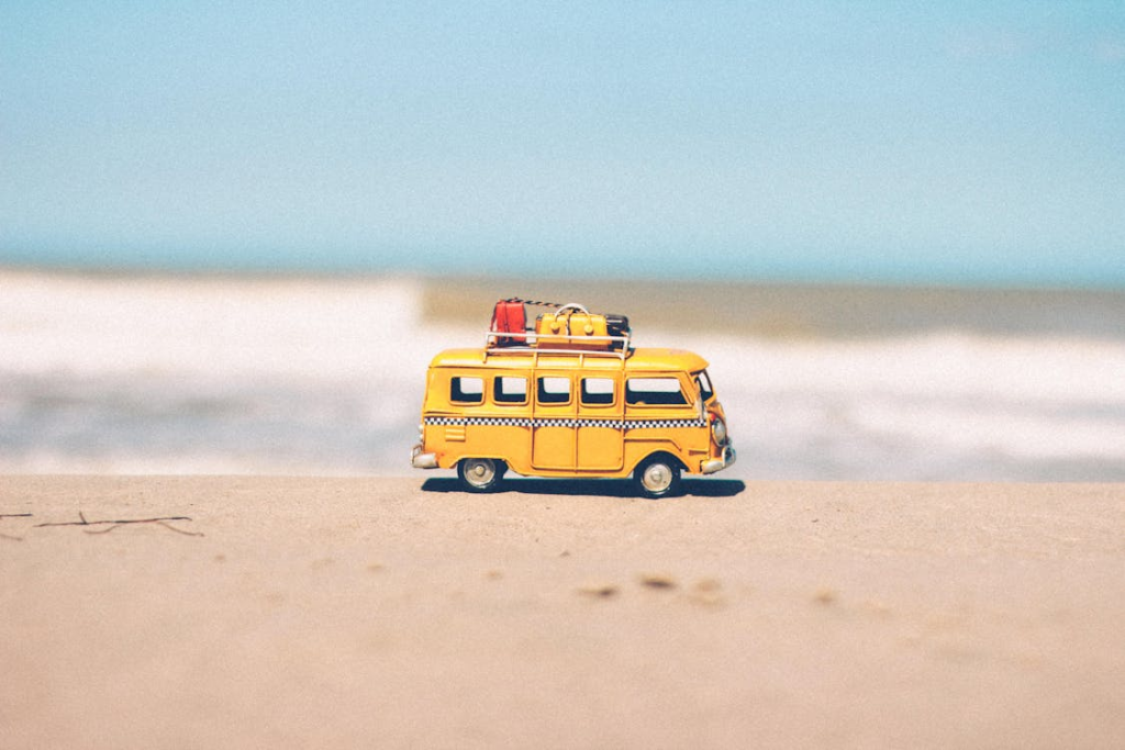 A toy yellow van with luggage on the roof, placed on a sandy surface with the sea in the background.