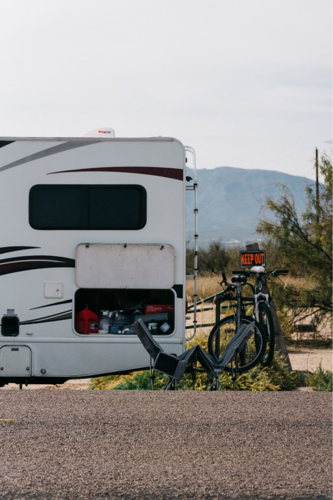 The rear side of a motorhome, with an open compartment showing items stored inside, bicycles mounted on the back, and camp chairs folded nearby.