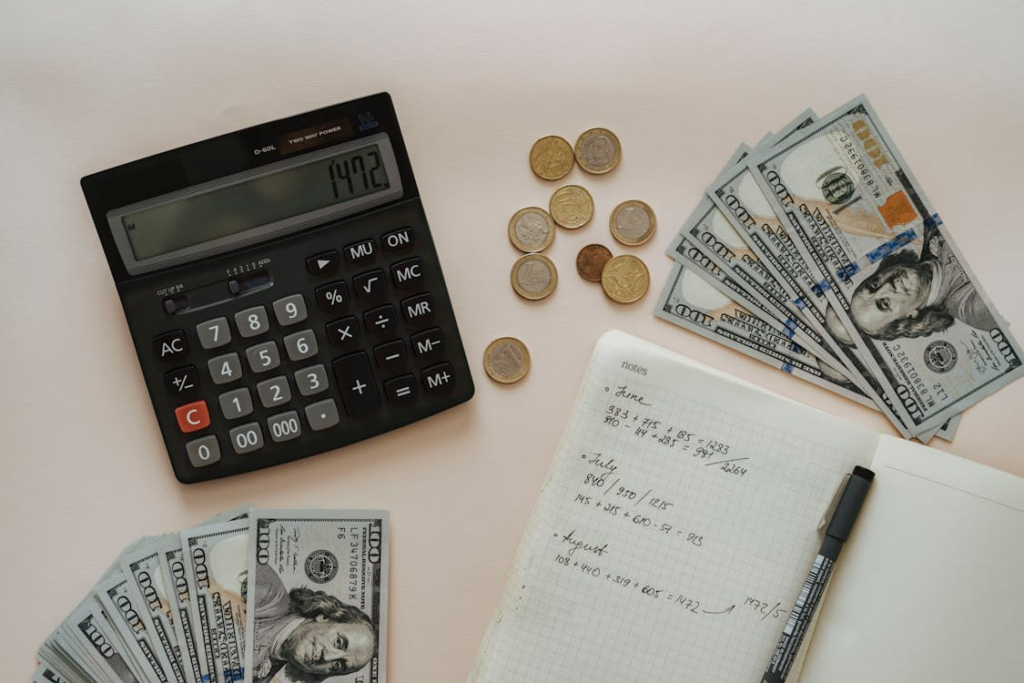 A calculator, coins, paper bills, and a notebook with handwritten financial notes arranged on a white surface.