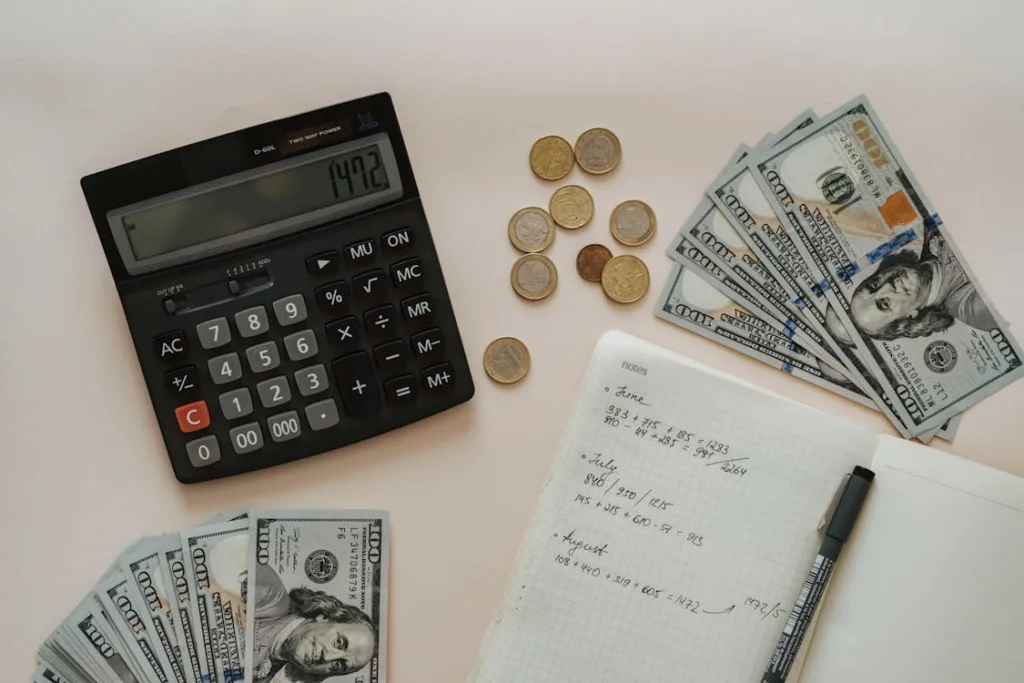 A calculator, coins, paper bills, and a notebook with handwritten financial notes arranged on a white surface.