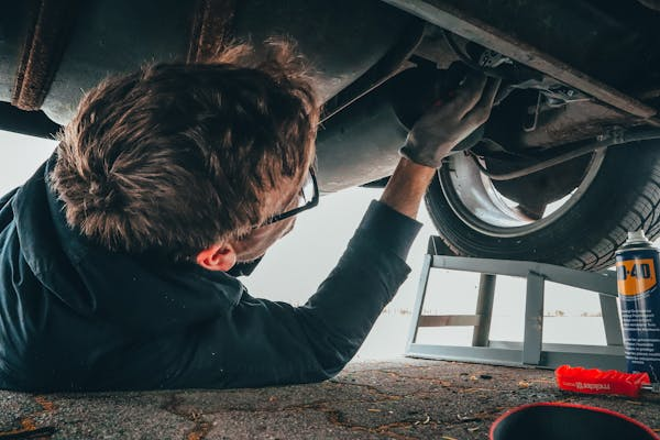 A man working under a vehicle, possibly making repairs, with various tools and a can of WD-40 nearby.