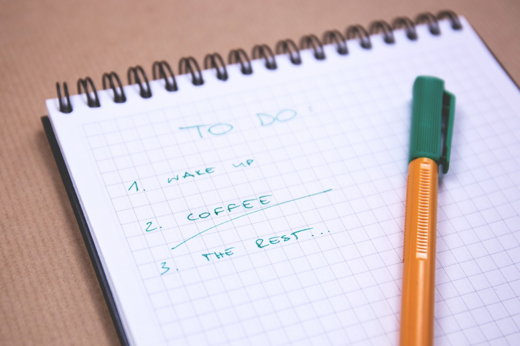 A to-do list on a spiral notebook with tasks "Wake up," "Coffee," and "The rest" checked off, with a pen beside it.