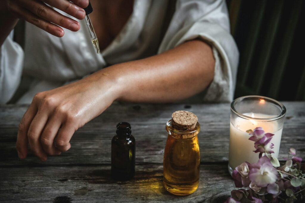 A woman applying oil to their arm with a dropper, next to bottles of oil and a lit candle on a wooden table.