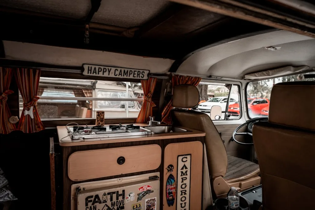 The interior of a camper van with a sign that reads "HAPPY CAMPERS," a small kitchen area, and cozy orange curtains.