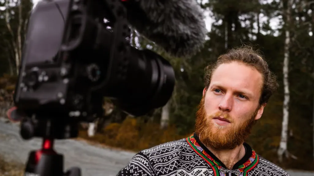 A man with a beard and long hair standing in an outdoor setting, talking to a camera mounted on a tripod.