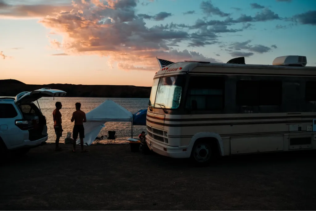 Two people standing and talking near a motorhome parked by the water at sunset, with a tent and an SUV nearby.