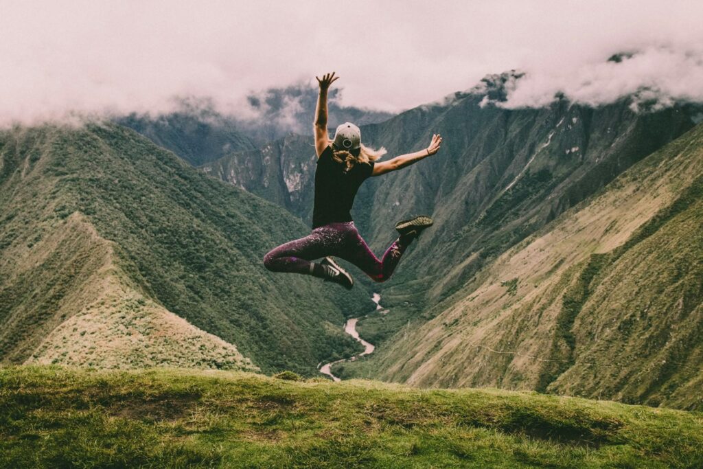 A woman in mid-air, joyfully leaping with raised arms, overlooking a scenic mountain valley.