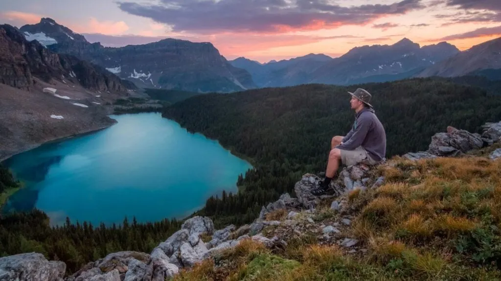 A man sitting on a rocky cliff overlooking a serene, turquoise lake surrounded by forested mountains during sunset.