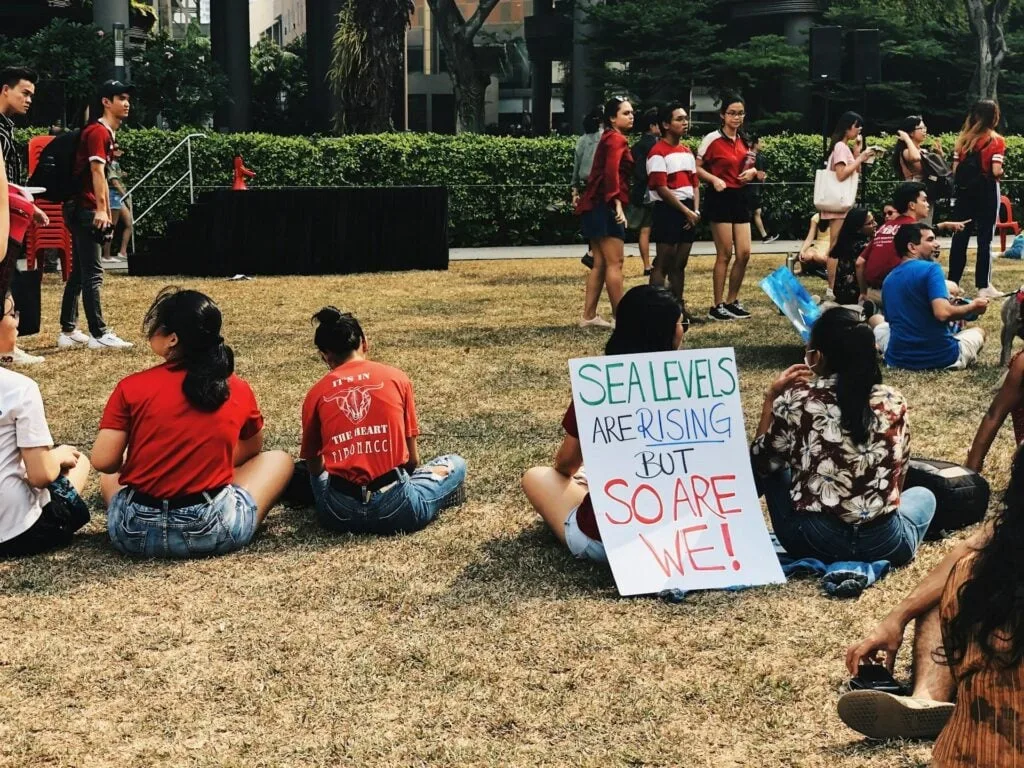 A group of people sitting on the grass at a gathering, with a sign that reads “Sea levels are rising but so are we!” in the foreground.