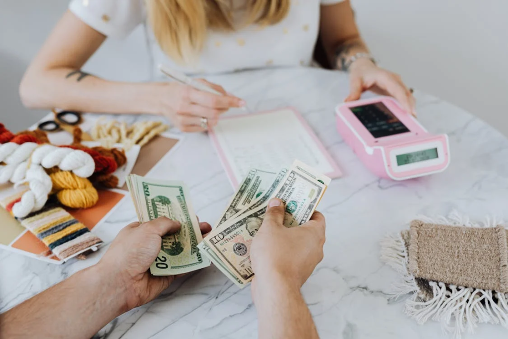 Two people at a table with one counting US dollar bills and the other writing on a notepad, with a payment terminal and yarn in the background.