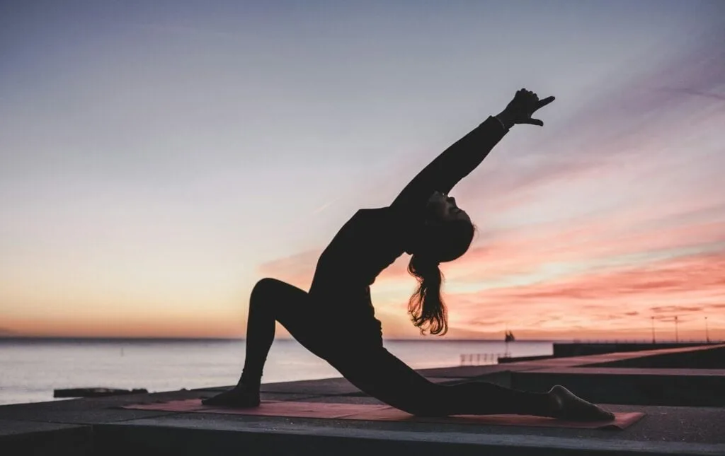 A person performing a yoga pose outdoors at sunset, with a colorful sky and the ocean in the background.