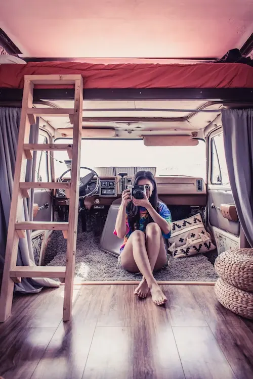 A woman sitting on the floor of a camper van, taking a photo with a camera, with a ladder leading to a lofted bed visible.