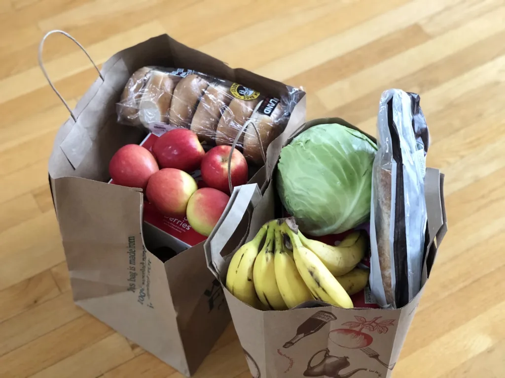 Two paper grocery bags filled with various fruits, vegetables, and bread, placed on a wooden floor.