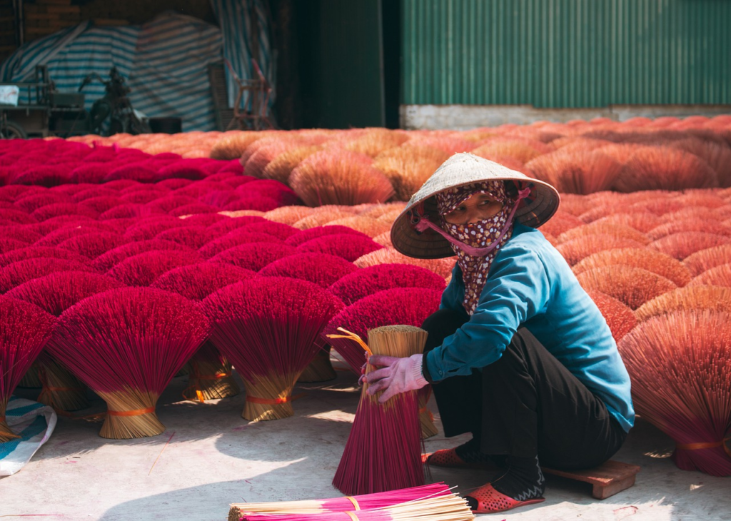 A worker wearing a hat and face covering, sitting among bundles of drying incense sticks in an outdoor setting.