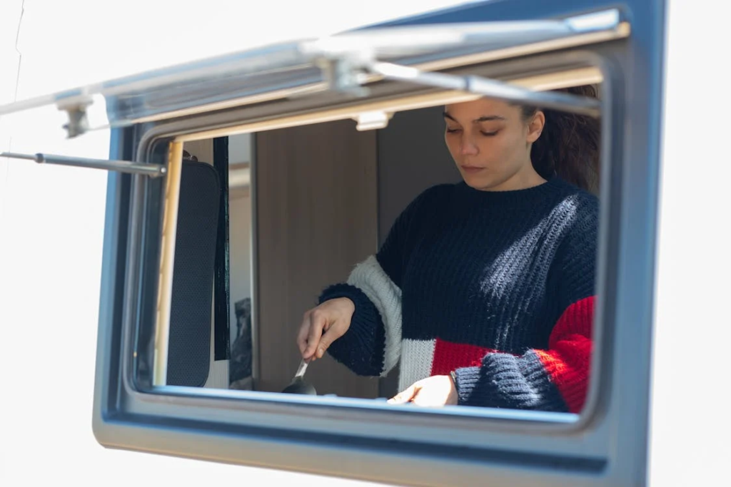 A woman cooking inside a camper van, viewed through an open window.