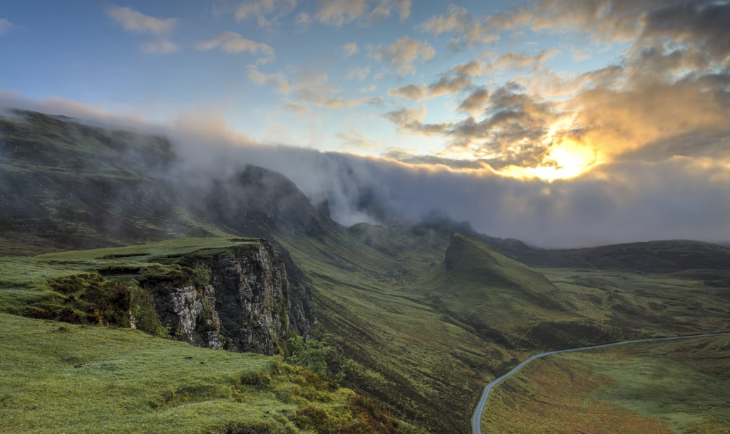  A scenic view of a sunrise or sunset over a hilly landscape with mist and a winding road.