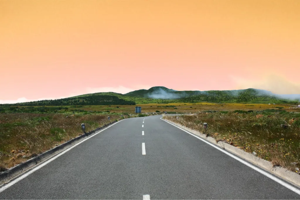 An empty road stretching towards green hills with an orange sky in the background.