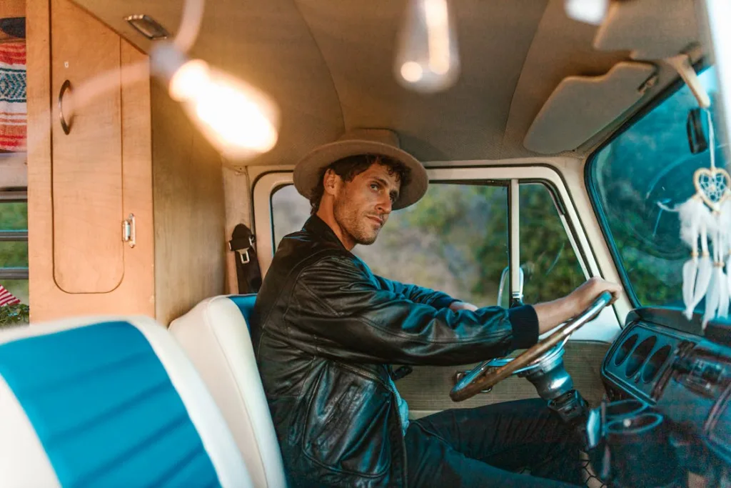 A man in a hat and leather jacket sits behind the wheel inside a retro campervan, looking contemplative.