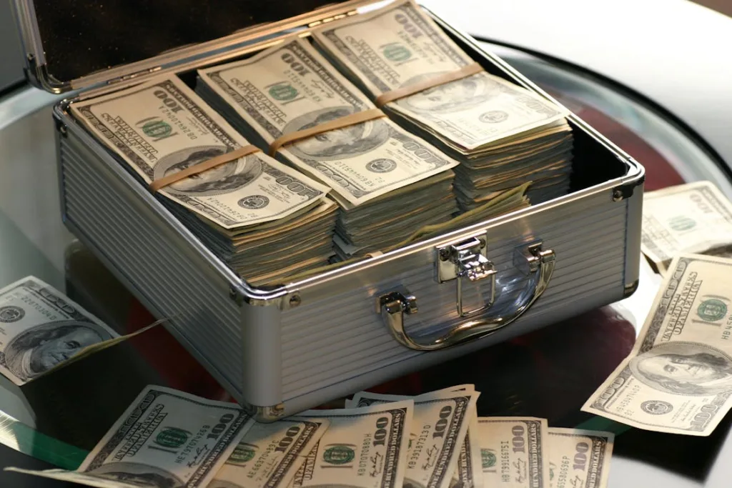 A metal briefcase filled with stacks of US dollar bills, with additional bundles of cash scattered around it on a glass table.