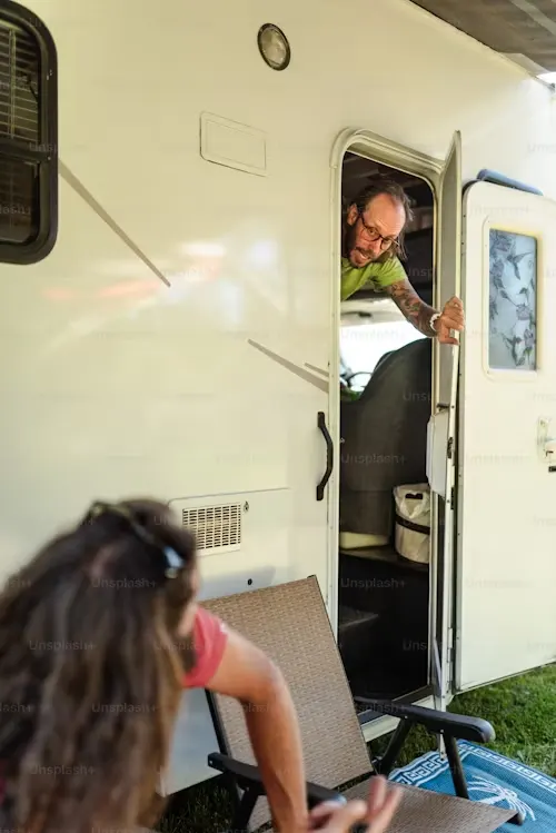 A person leaning out of an RV door, conversing with someone seated on a chair outside.