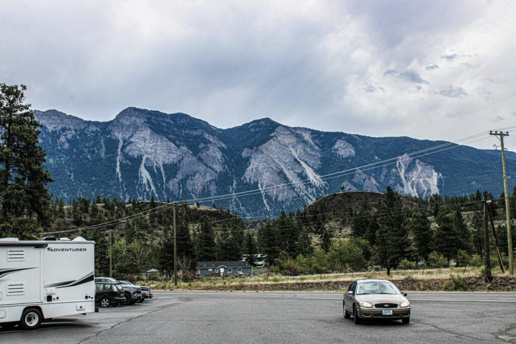 A parking lot with an RV and several cars, set against a backdrop of tall mountains under a cloudy sky.