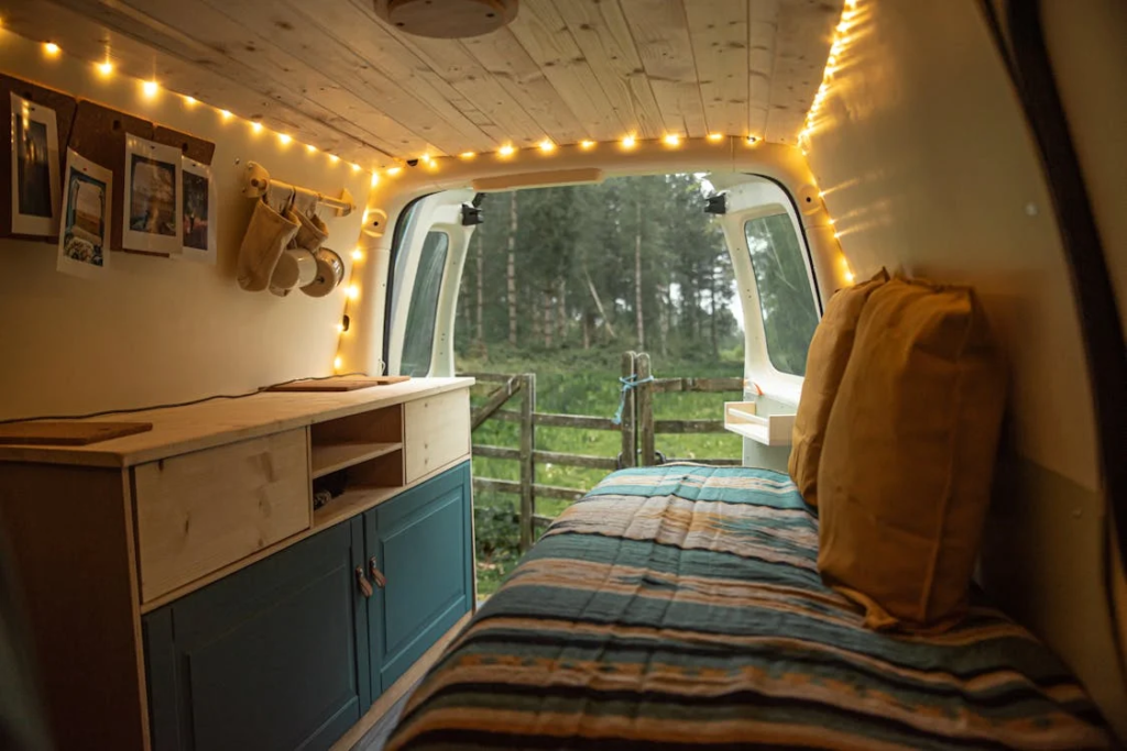 Interior of a camper van decorated with string lights and featuring a bed and cabinets, overlooking an outdoor landscape.