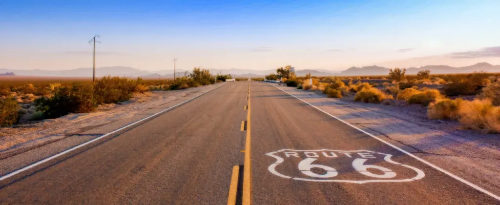 A straight, open road with "Route 66" marked on the pavement, extending into the horizon with a sunset sky and desert landscape.