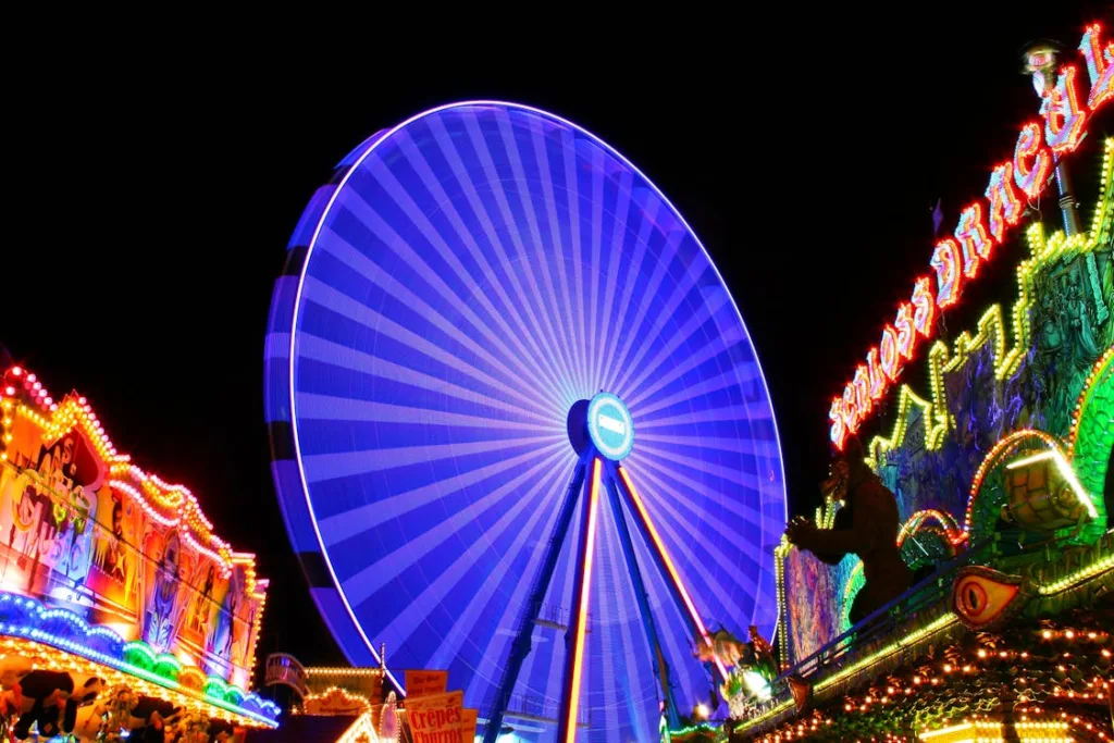 A vibrant night scene at a carnival featuring a large, illuminated Ferris wheel and colorful, lit-up attractions and signs.