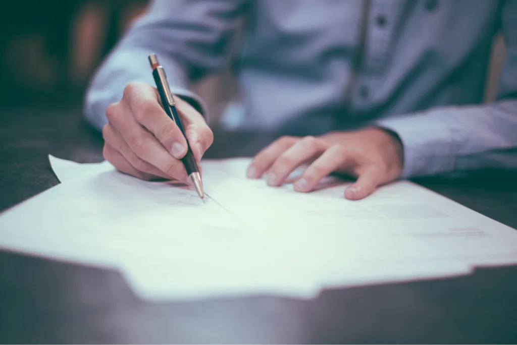 A person writing on documents with a pen on a table.