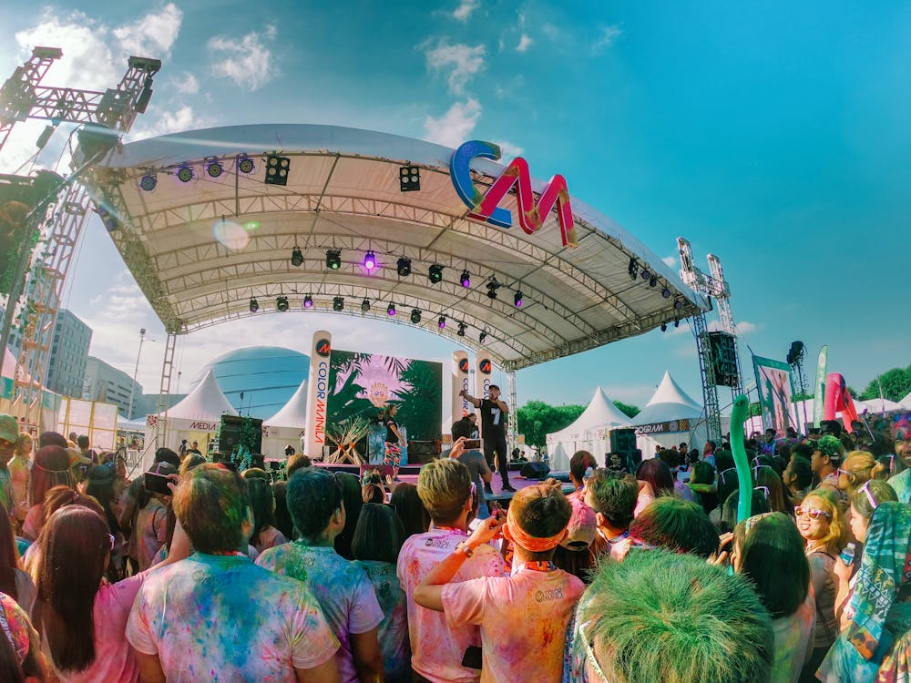 A large crowd of people attending an outdoor concert or event with a stage under a white canopy, colorful lighting, and a large logo.