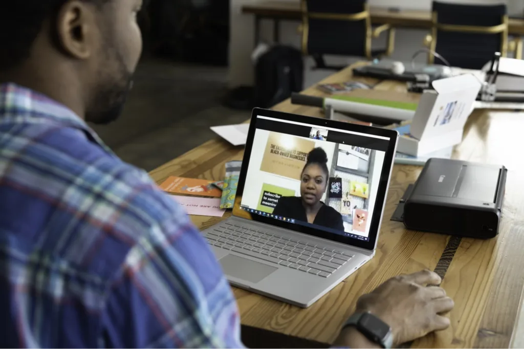 A man participating in a video call on a laptop, sitting at a table with various papers and a printer around.