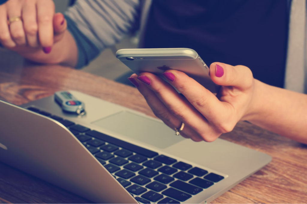 A close-up of someone’s hands; one is holding a smartphone, and the other rests on the table with a laptop and USB drive.