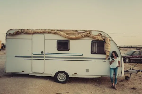 A person leaning against a white travel trailer with rolled-up canvas on the roof, holding a phone.