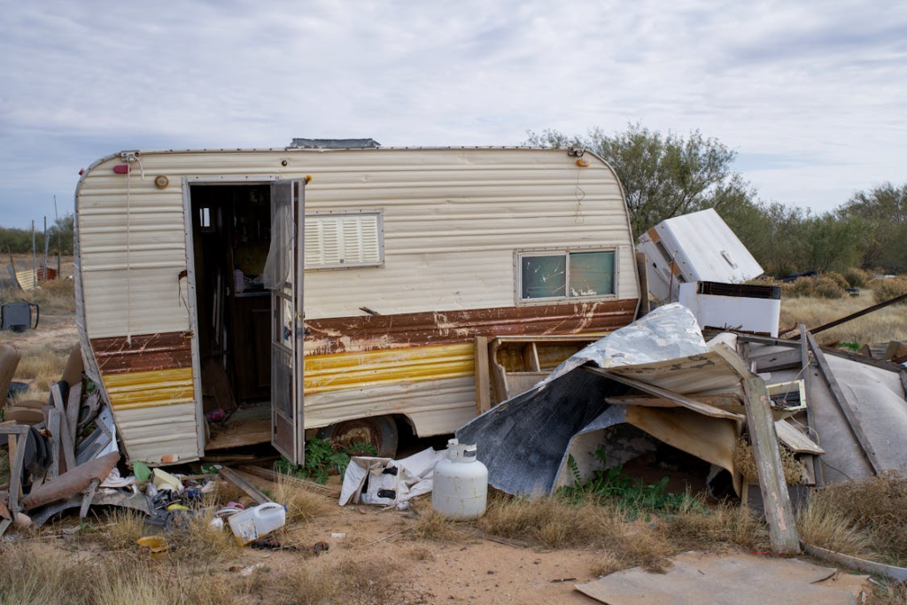 An old, weathered camper parked in a cluttered area with various discarded items around it.