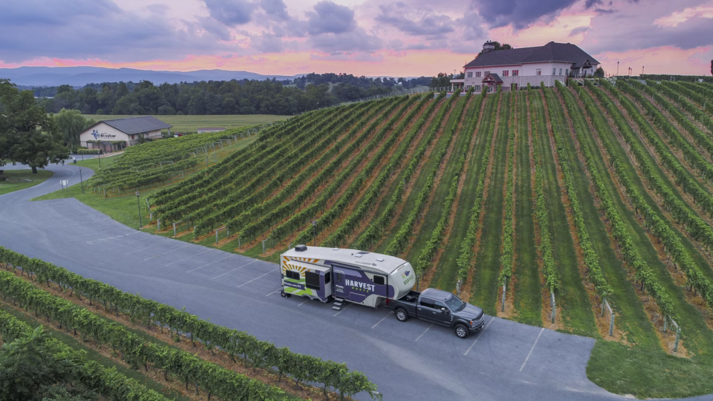 A large camper trailer parked at a vineyard, with rows of grapevines and a large building on a hill at sunset.