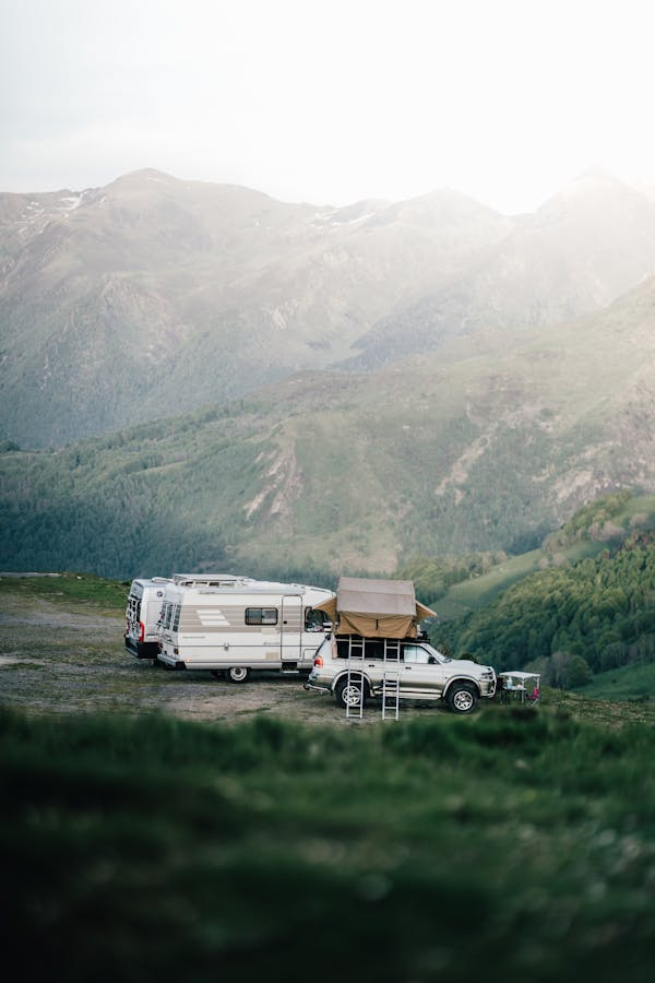 An off-road vehicle with a roof tent parked next to a travel trailer in a mountainous area under a cloudy sky.