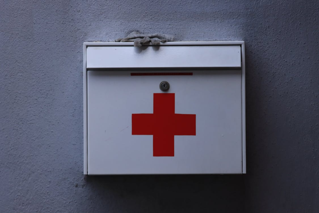 A white first aid kit box with a red cross drawn on it mounted on a gray wall.