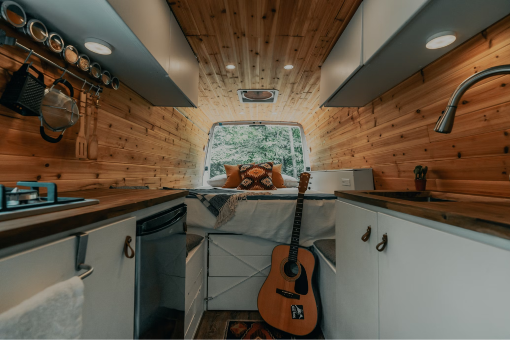 Interior of a rustic camper van featuring a wooden aesthetic with kitchen cabinets, a bed, and a guitar placed nearby.