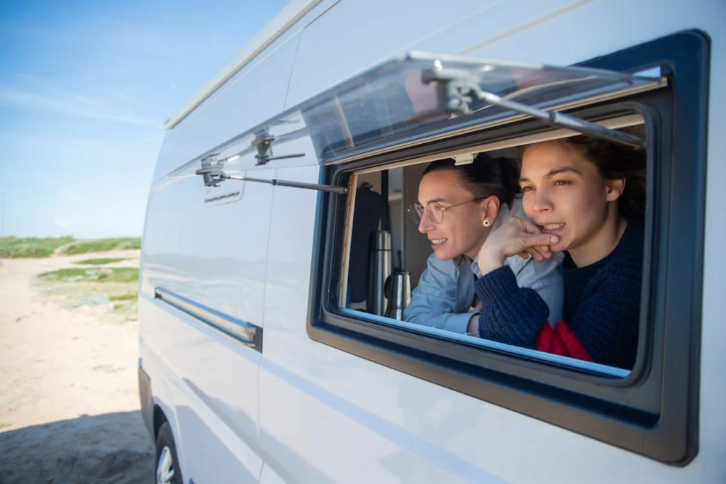 Two people leaning out of the side window of a white camper van, enjoying the outdoors on a sunny day.