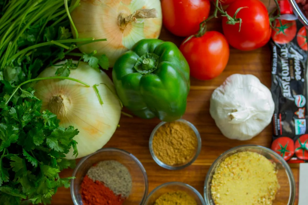 Fresh vegetables and ingredients arranged on a wooden surface, including onions, green bell peppers, tomatoes, garlic, and various spices in small glass bowls.