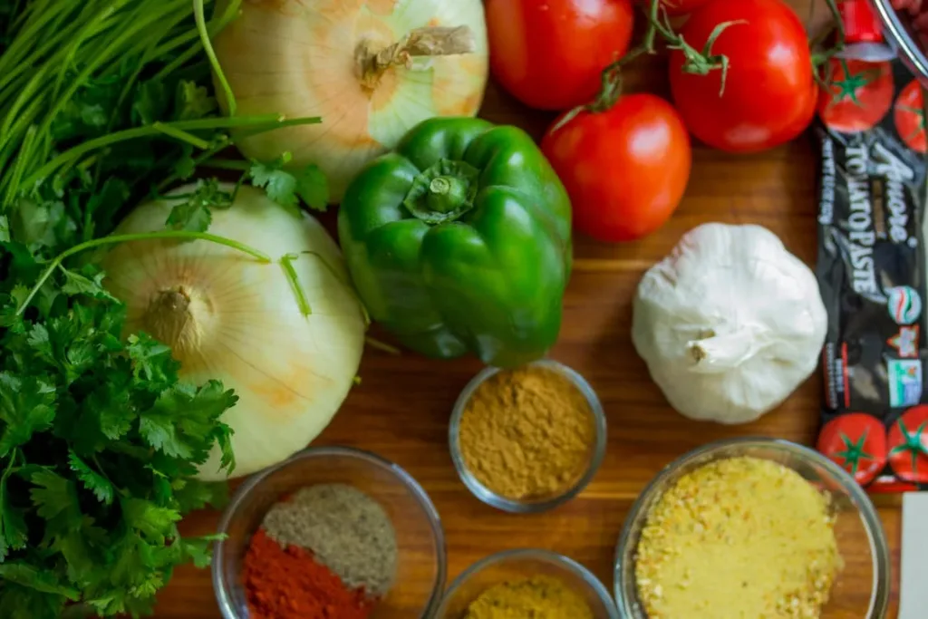 Fresh vegetables and ingredients arranged on a wooden surface, including onions, green bell peppers, tomatoes, garlic, and various spices in small glass bowls.