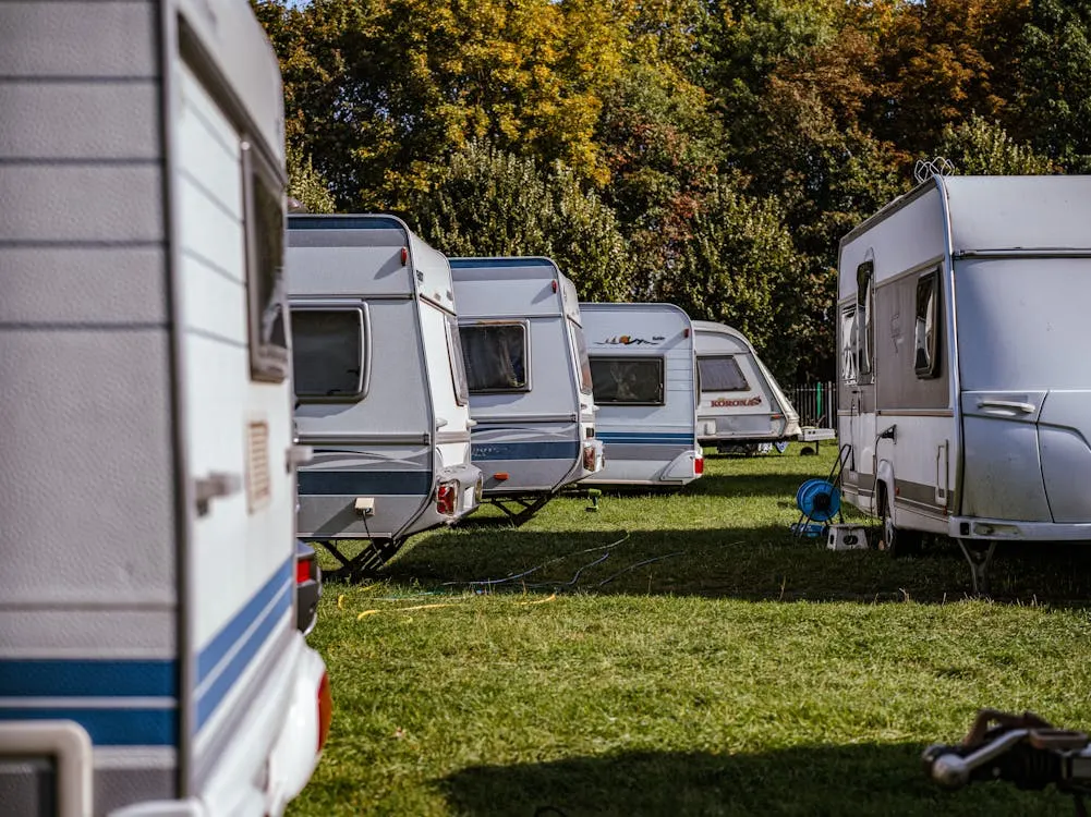 A row of white and blue caravans parked on green grass with trees in the background.