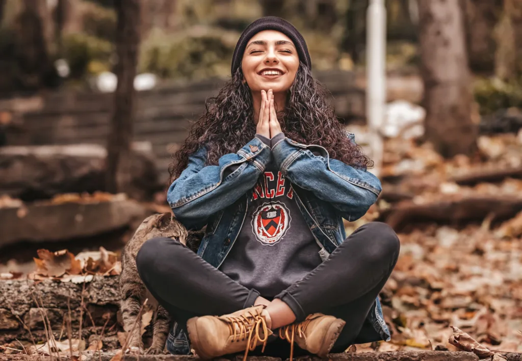 A woman sitting cross-legged outdoors, smiling with their hands pressed together in front of them, surrounded by autumn leaves.