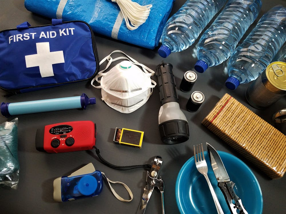 A spread of various emergency and survival supplies, including a first aid kit, water bottles, masks, a flashlight, batteries, a radio, matches, a whistle, plates, cutlery, and packaged food.