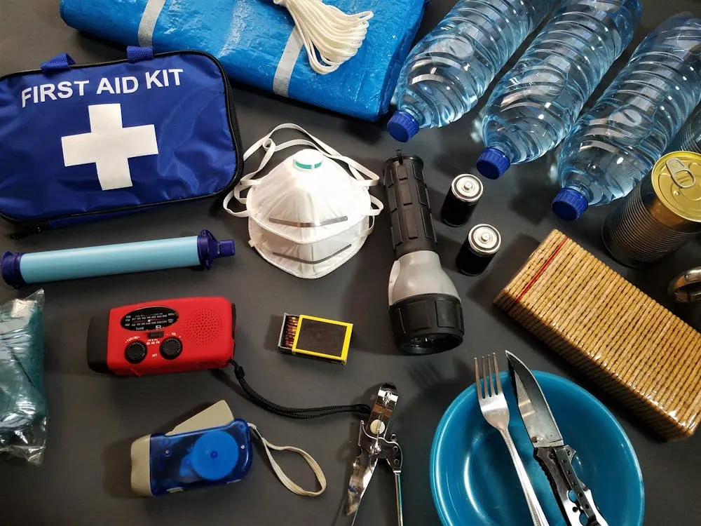 A spread of various emergency and survival supplies, including a first aid kit, water bottles, masks, a flashlight, batteries, a radio, matches, a whistle, plates, cutlery, and packaged food.