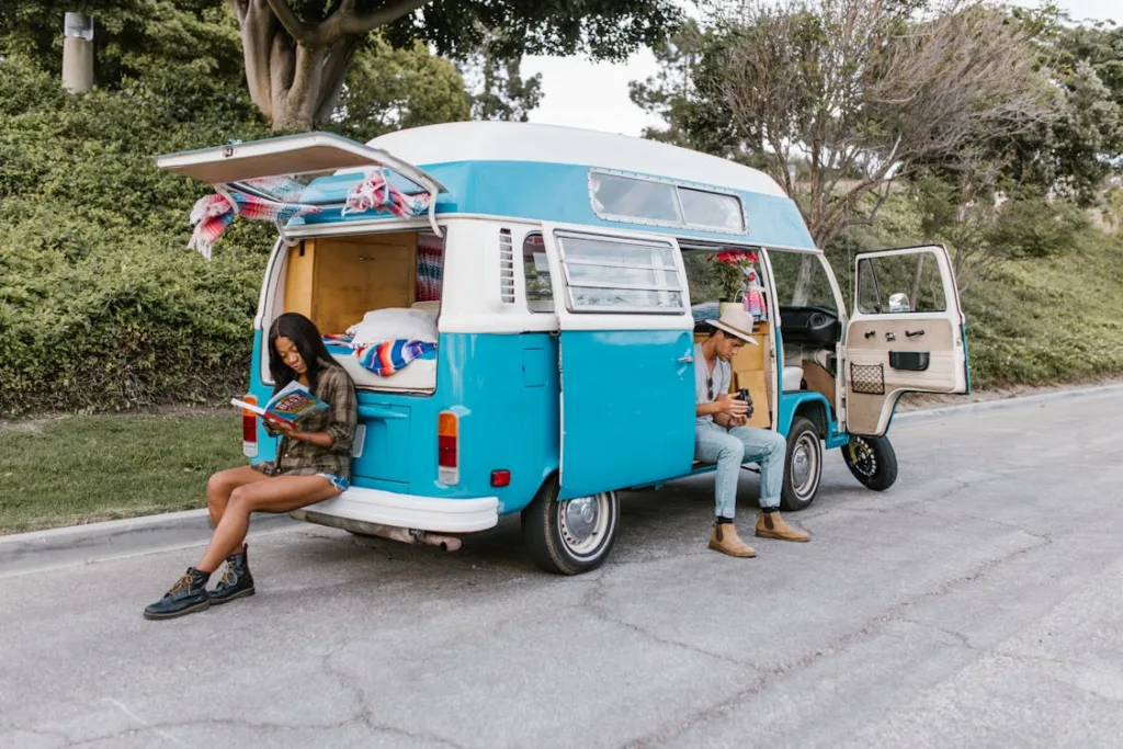  A woman sitting at the back of a blue and white van reading a book, and a man sitting at the side door, using a phone.