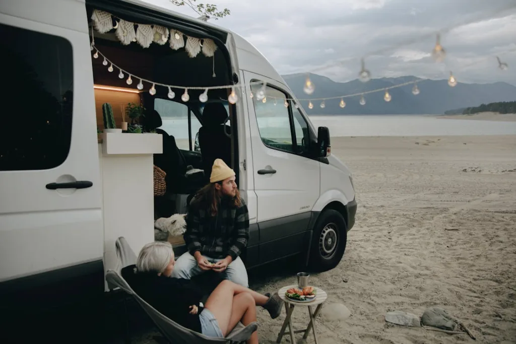 A couple sitting outside a white campervan parked on a beach, with string lights hanging above and a scenic view of mountains and water in the background.