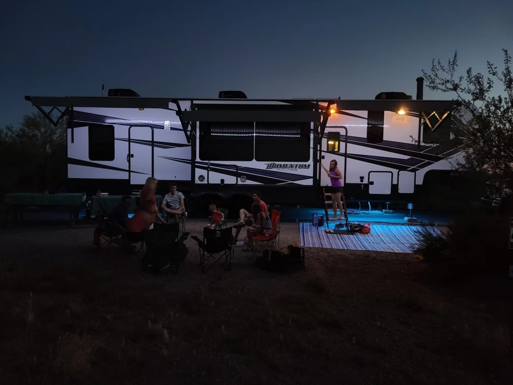 A group of people sitting and standing outside a large motorhome at night, with the motorhome's lights on.